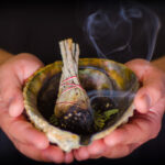 A picture of hands cradling an abalone shell with burning sage and a pleasant smoke billowing from the burnt ends of the sage